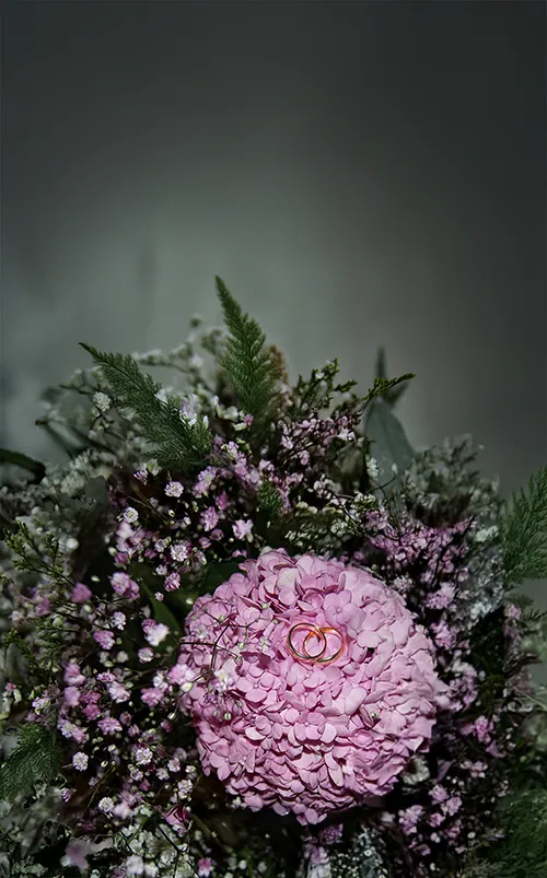 Wedding Photography in Landsberg – Close-up of wedding rings placed on a pink bouquet with white flowers and green leaves, set against a soft green-to-black gradient background.
