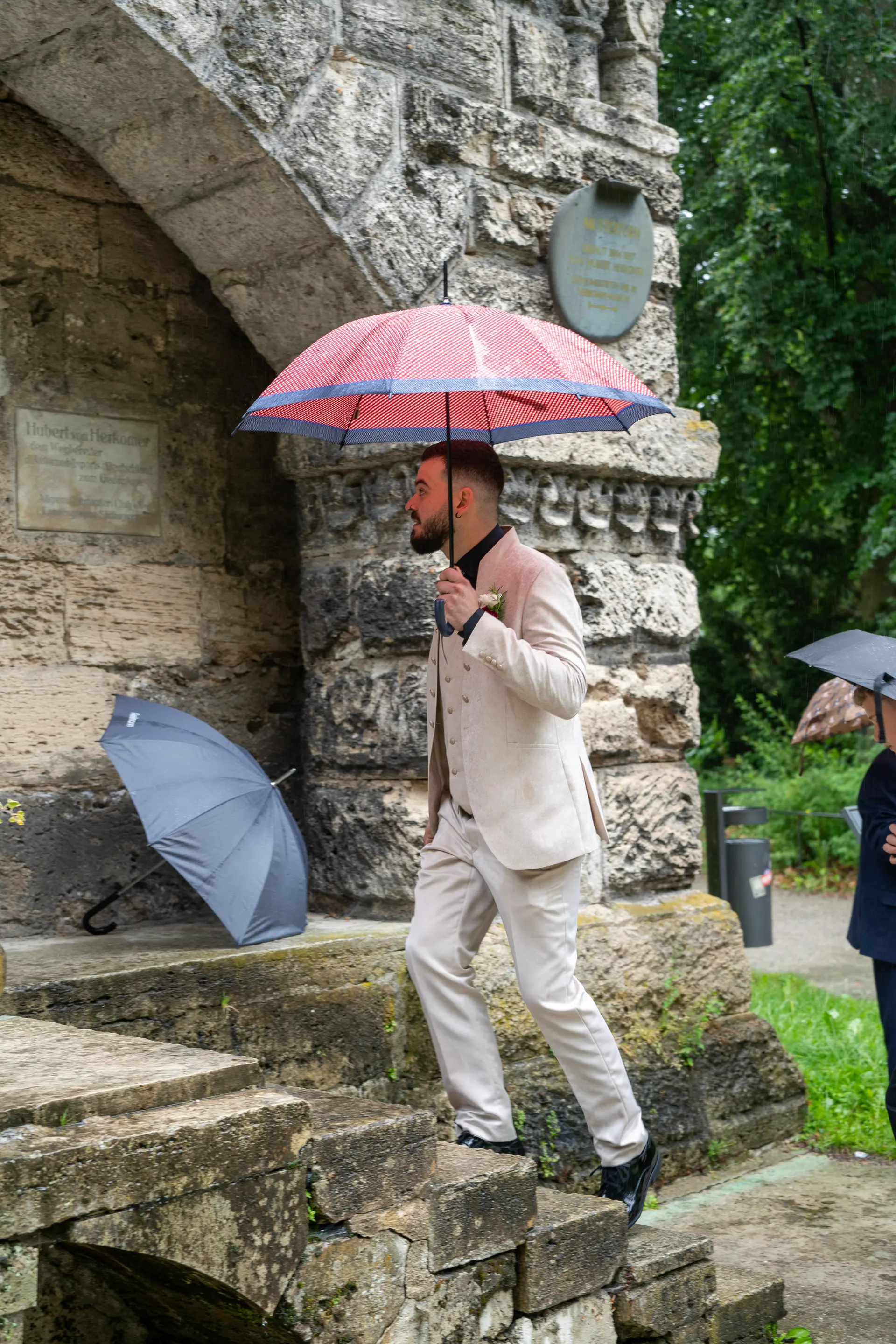 Groom in a light suit climbing the stone steps at the Mutterturm under a red umbrella