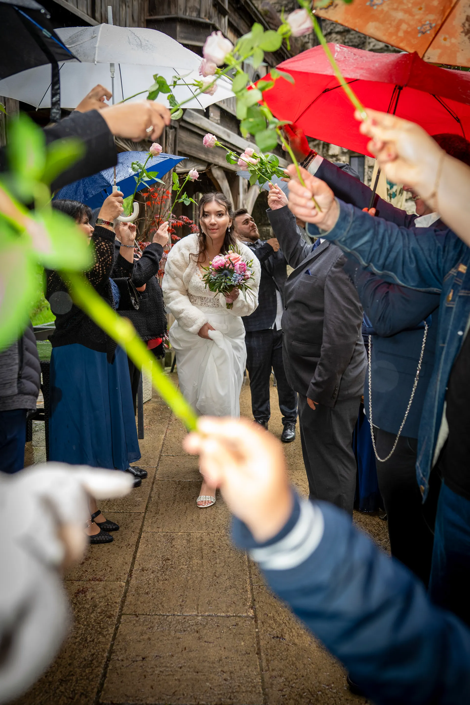 Guests form an arch of roses and umbrellas in the rain for the bride
